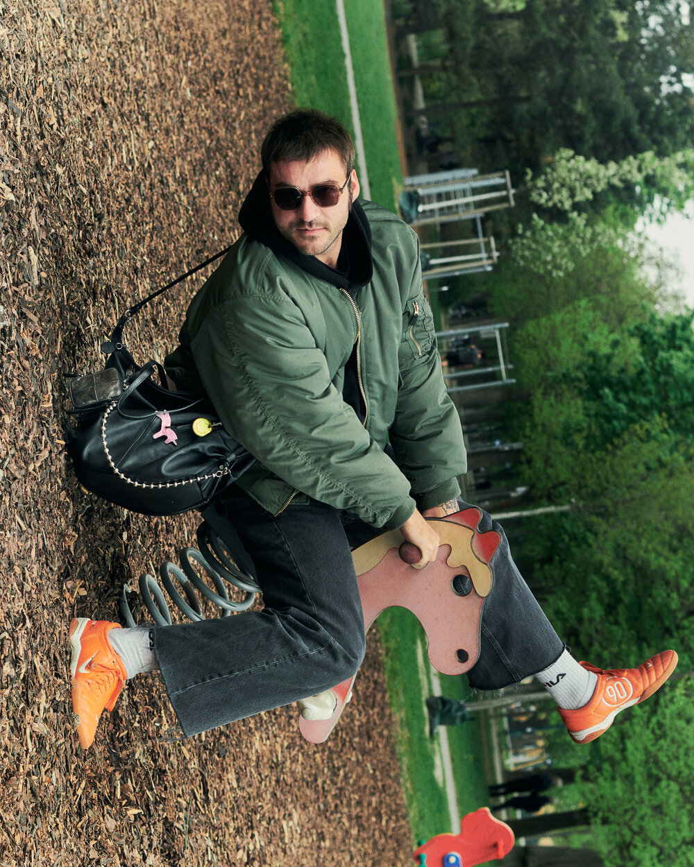 Ein Erwachsener mit grüner Jacke und orangefarbenen Turnschuhen sitzt auf einem Springreiter auf einem Spielplatz und hält eine schwarze INA KENT-Tasche. Im Hintergrund sind Bäume und Parkeinrichtungen zu sehen.