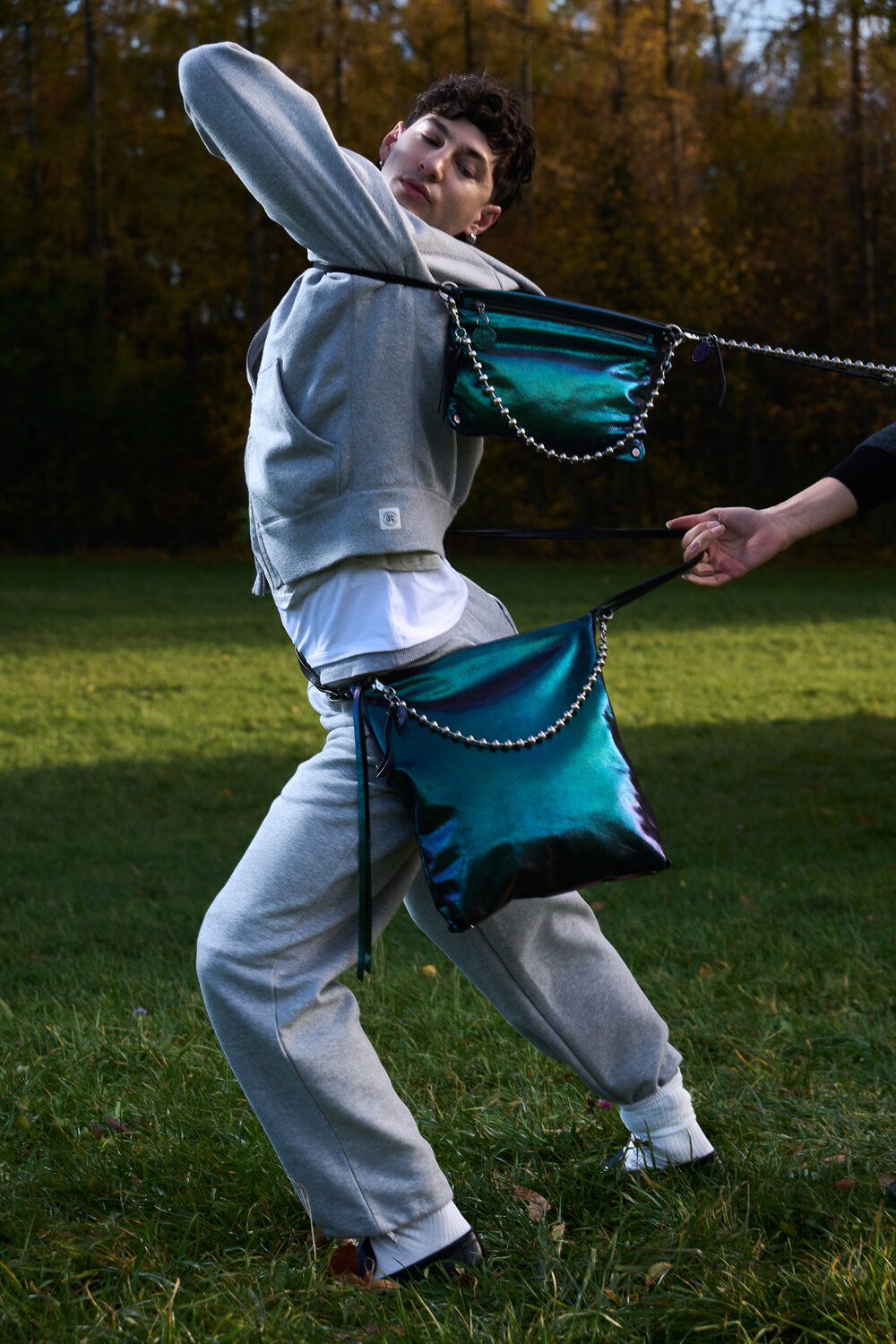 A person in a grey tracksuit poses outdoors while being tugged backward by two metallic teal INA KENT handbags with chain straps, held by someone out of frame.