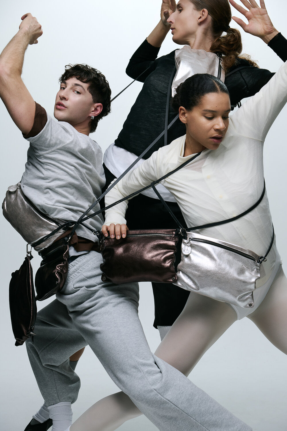 Three people pose dynamically, wearing neutral-toned outfits and INA KENT metallic bags connected by long straps that intertwine among them against a plain background.