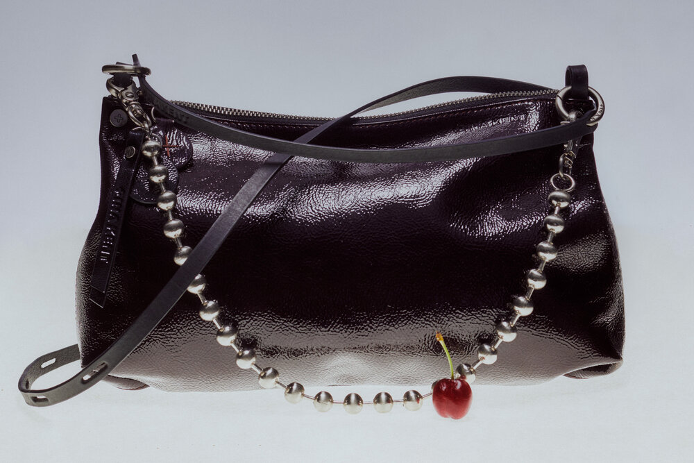 A shiny black leather INA KENT handbag with a beaded strap and a cherry attached to the bottom, displayed against a plain background.