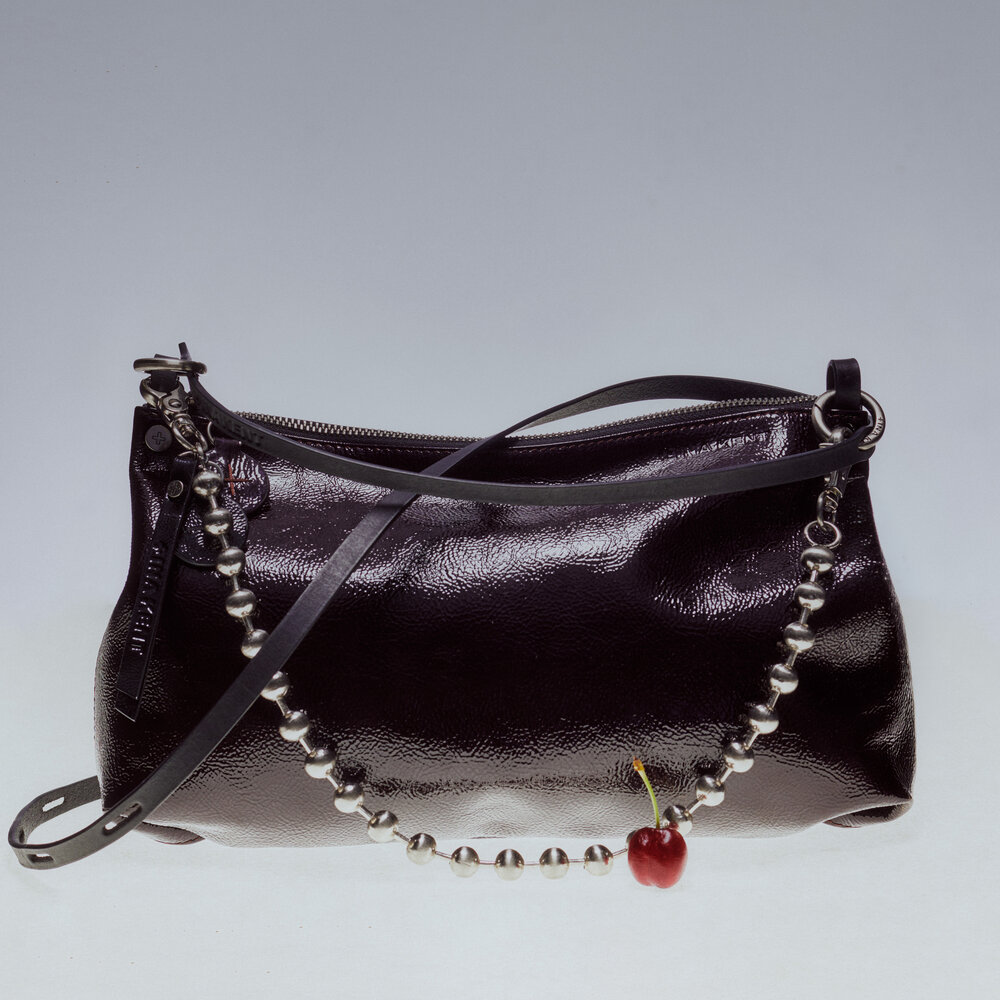 A shiny black leather INA KENT handbag with a strap decorated with silver beads and a single red cherry charm, set against a plain background.