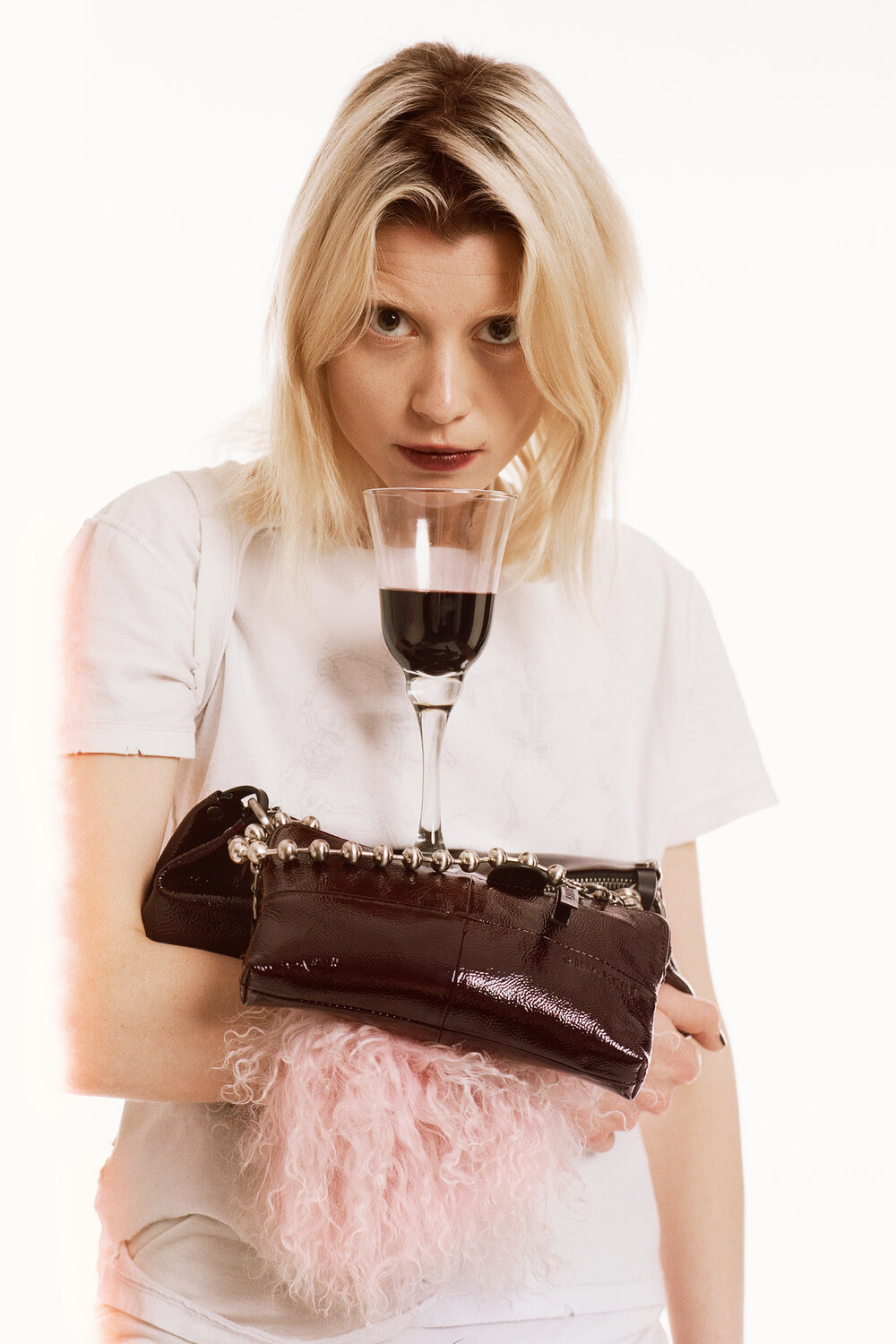 Eine Frau in einem weißen T-Shirt hält eine schwarze Handtasche von INA KENT und balanciert ein Glas Rotwein auf ihrem Unterarm, während sie direkt in die Kamera blickt.