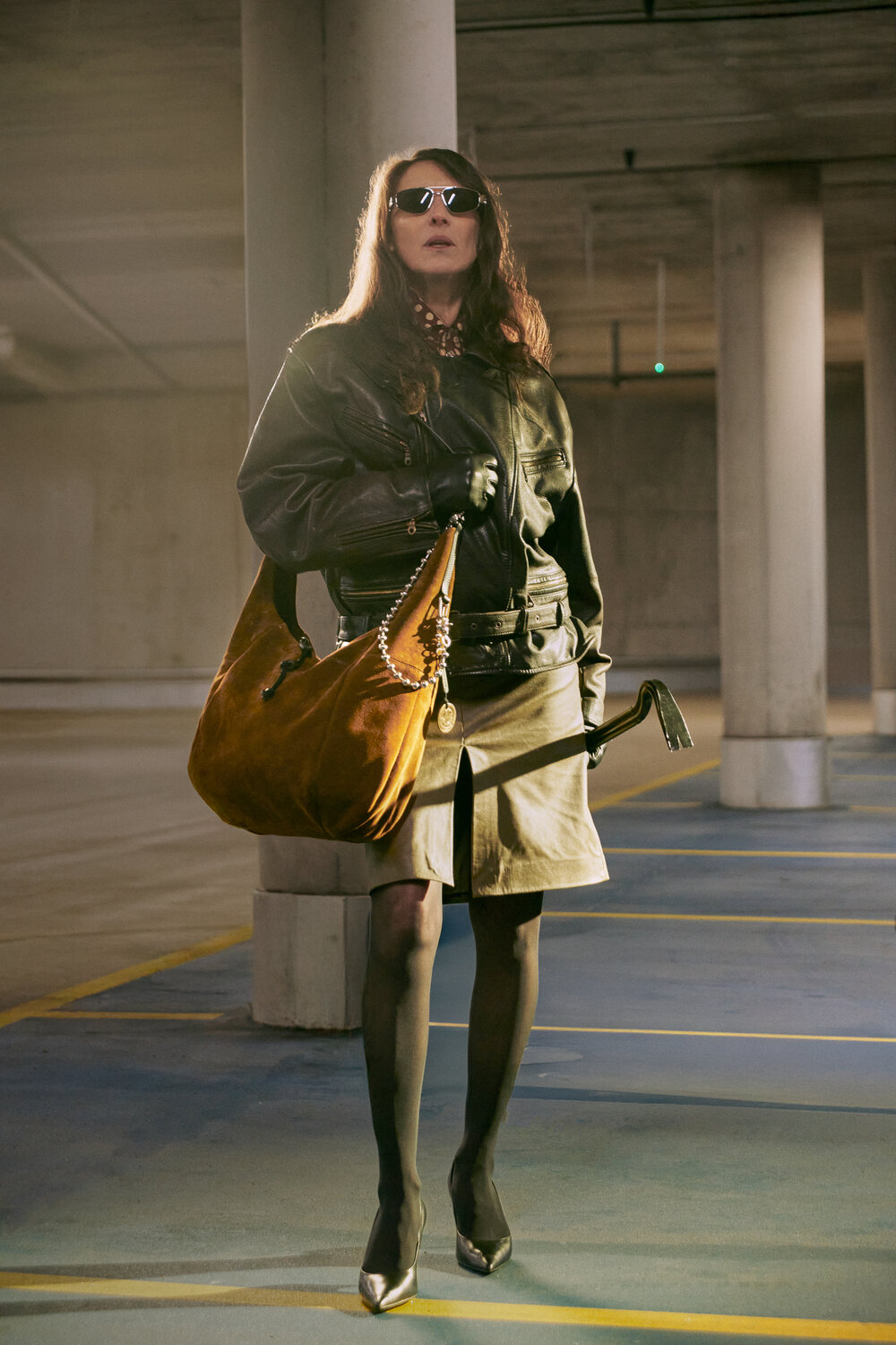 A person wearing sunglasses, a black leather jacket, a khaki skirt, and heels stands in a parking garage, holding a large brown INA KENT bag.