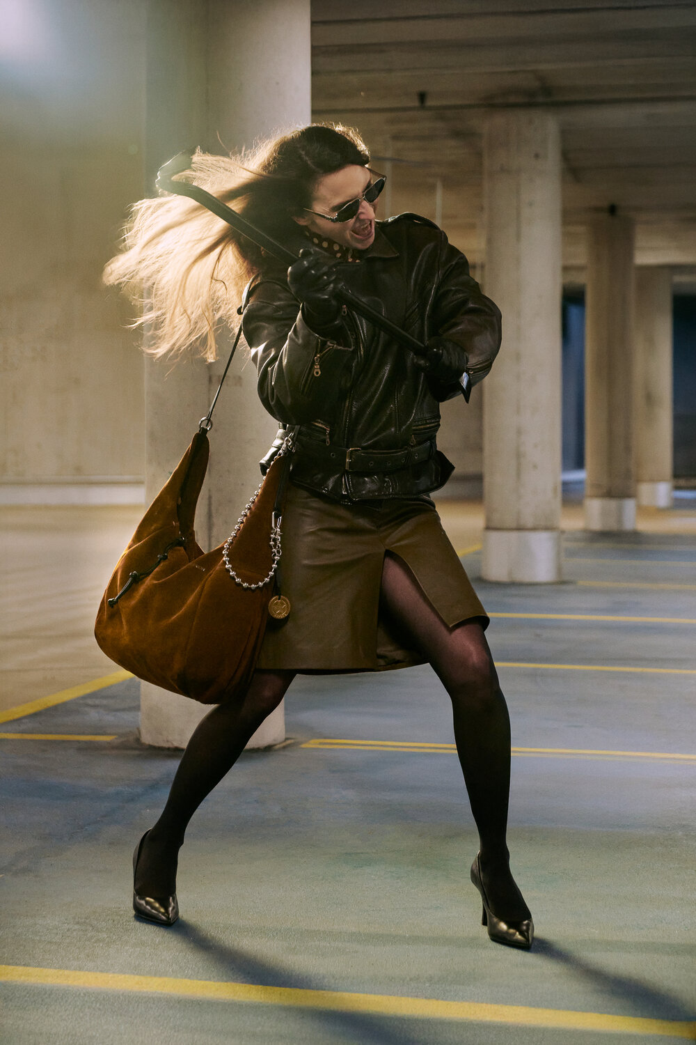 A woman in a leather jacket, skirt, and sunglasses swings a large brown INA KENT bag in an industrial parking garage.