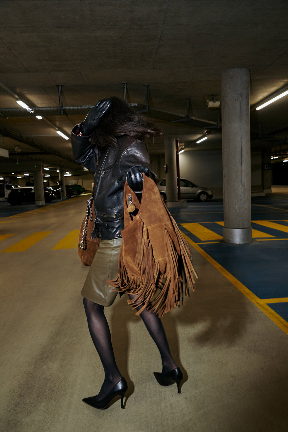 Person in a parking garage wearing a leather jacket, khaki skirt, black heels, and black gloves, carrying a large brown fringed INA KENT handbag while turning away from the camera.