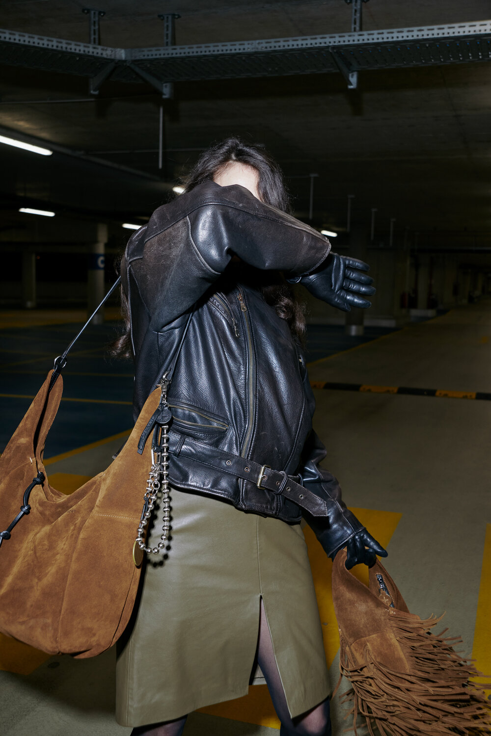 A person in a leather jacket and gloves covers their face with their arm while standing in a parking garage, carrying a large brown suede INA KENT bag and a fringed accessory.