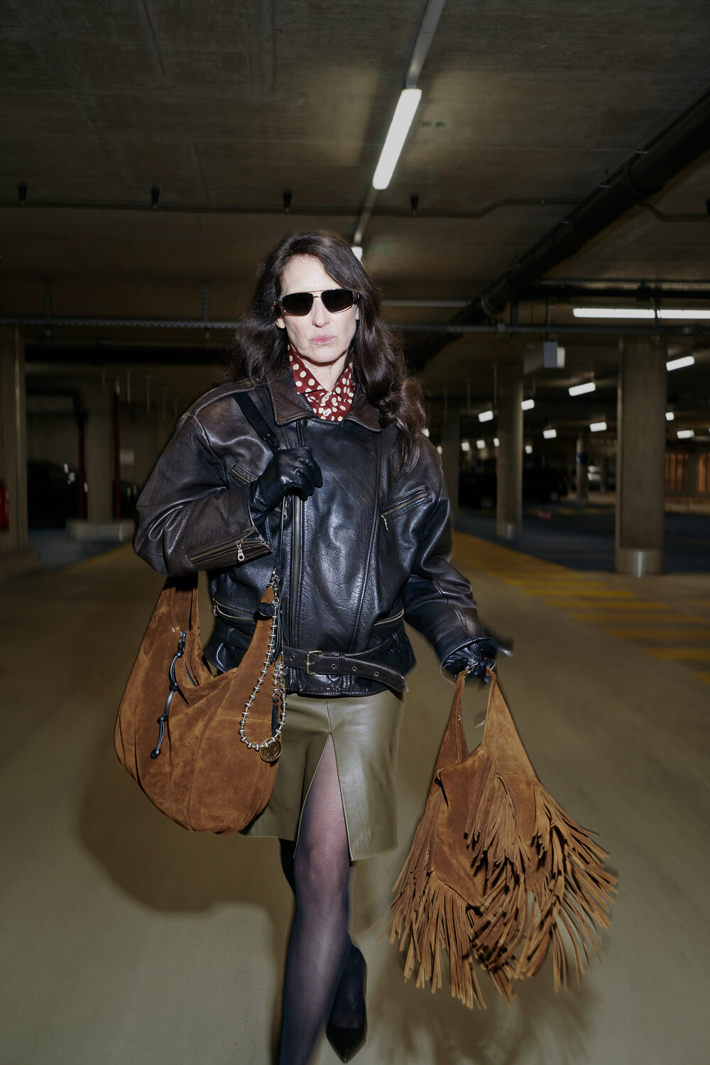 A woman wearing sunglasses, a leather jacket, gloves, and a patterned scarf walks in a parking garage carrying two brown fringed INA KENT bags.