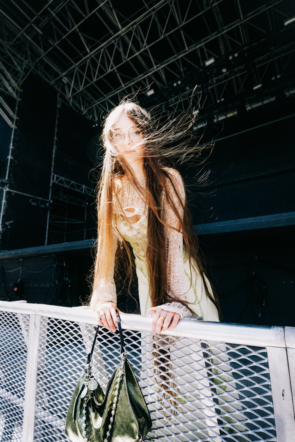 A person with long hair stands behind a metal barrier on an outdoor stage, holding onto the railing, with an INA KENT green handbag hanging from it.