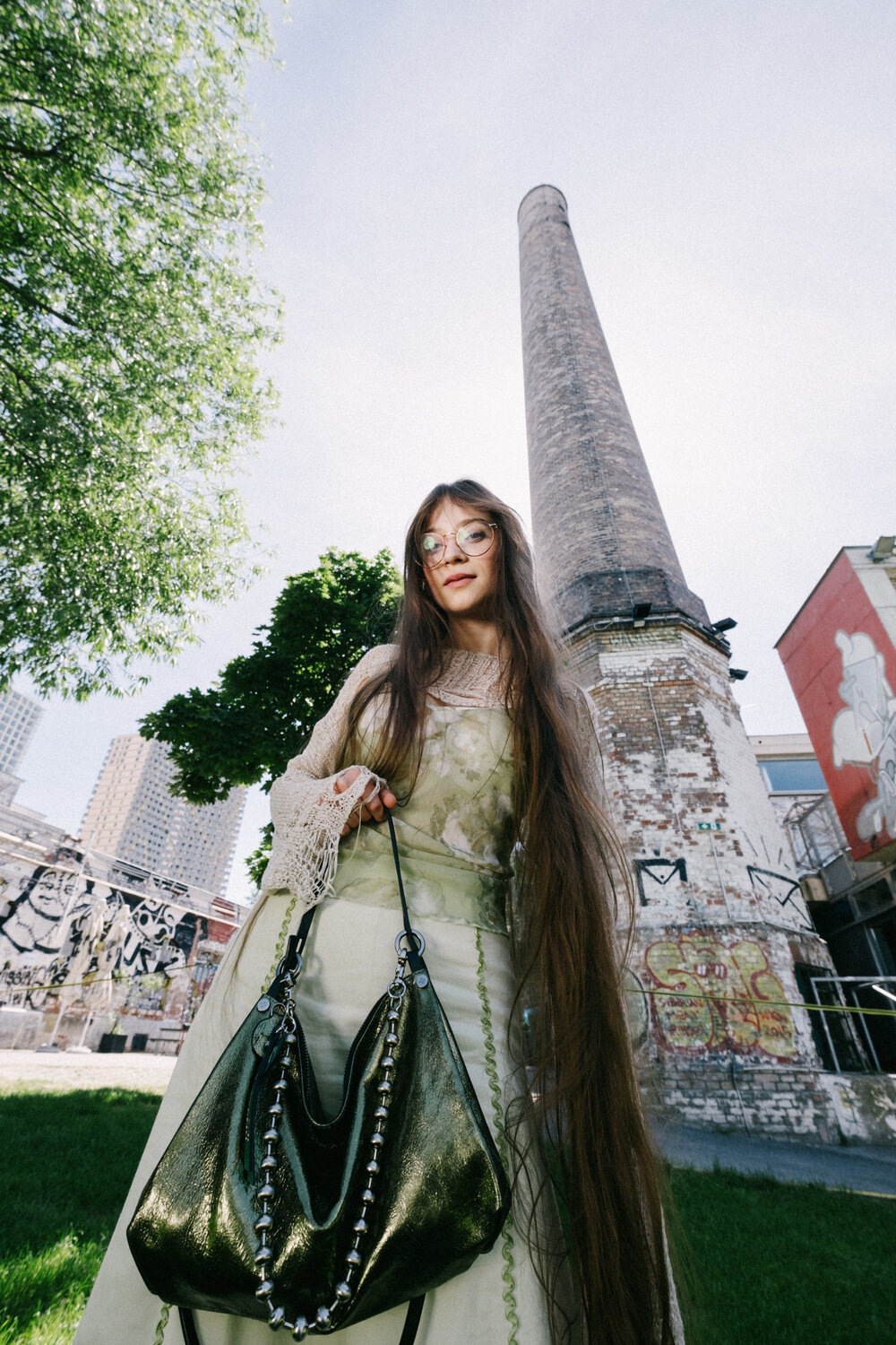 A woman with long hair and glasses stands outdoors in front of a tall brick smokestack, holding a metallic INA KENT handbag. Urban graffiti and city buildings are visible in the background.