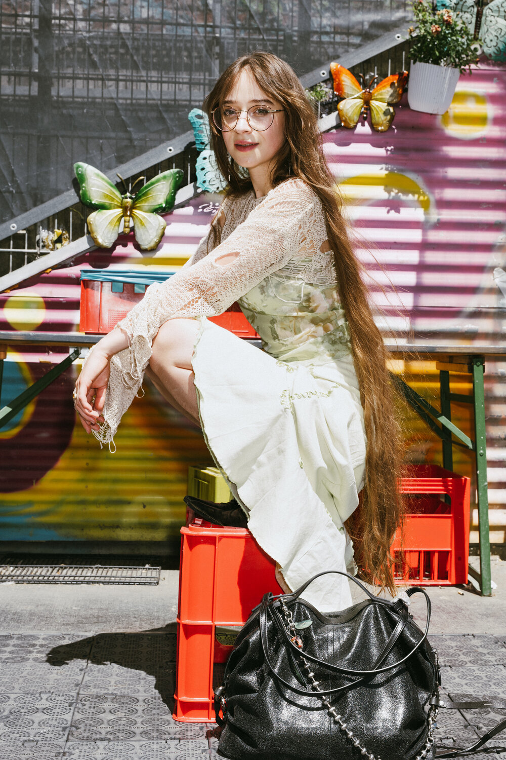 A woman with long brown hair and glasses sits on a red crate in front of a colorful mural with butterflies, wearing a light dress and lace top. A large black INA KENT bag rests on the ground beside her.