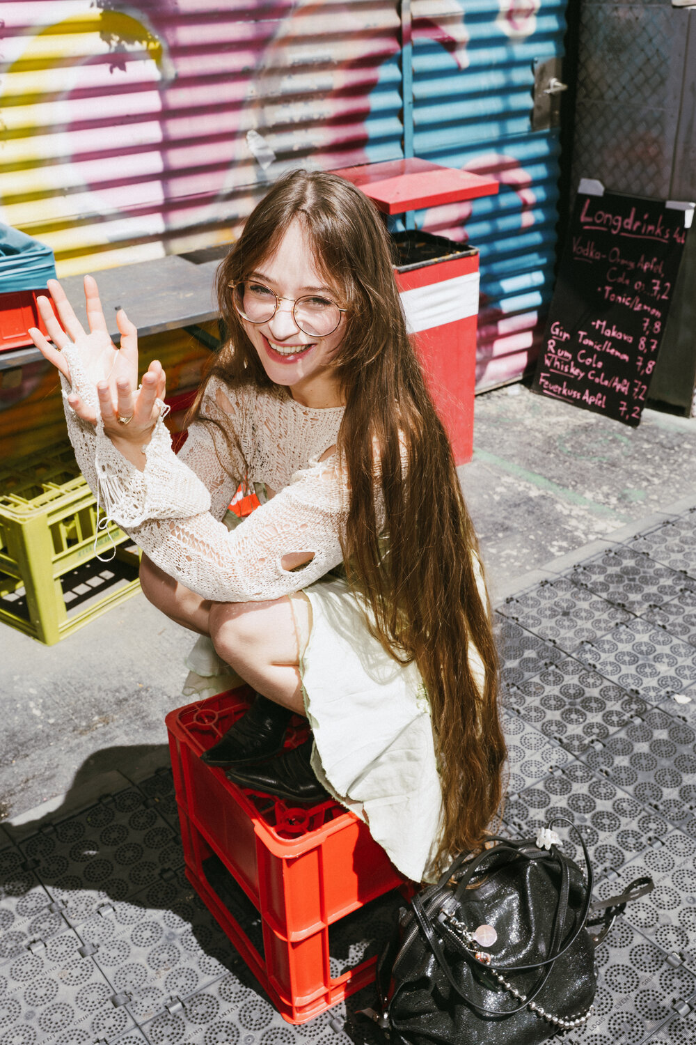 A woman with long hair and glasses squats on a red crate, smiling and waving outdoors next to a colorful wall, a menu board, and her stylish INA KENT bag.