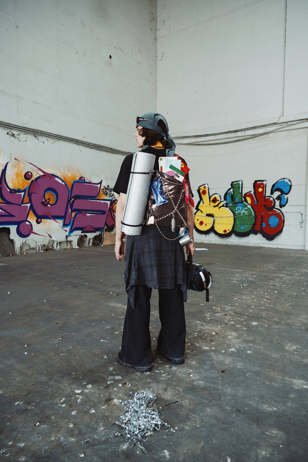 A person wearing dark clothing and carrying an INA KENT bag along with various camping gear stands in an empty warehouse with colorful graffiti on the walls.