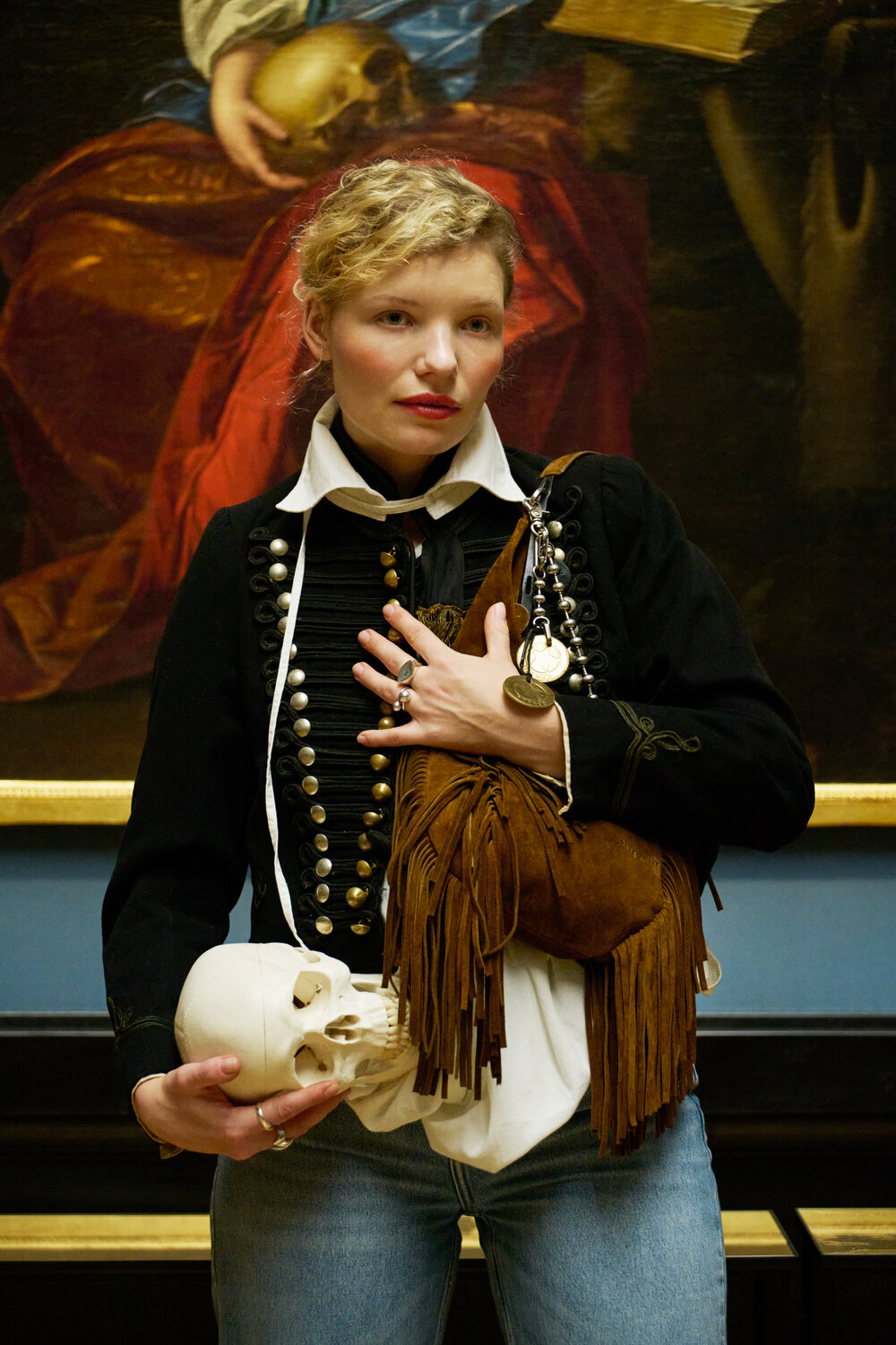 Eine Frau in einer schwarz-weißen Jacke hält vor einem klassischen Gemälde eine braune Tasche mit Fransen von INA KENT und einen weißen Totenkopf in der Hand.