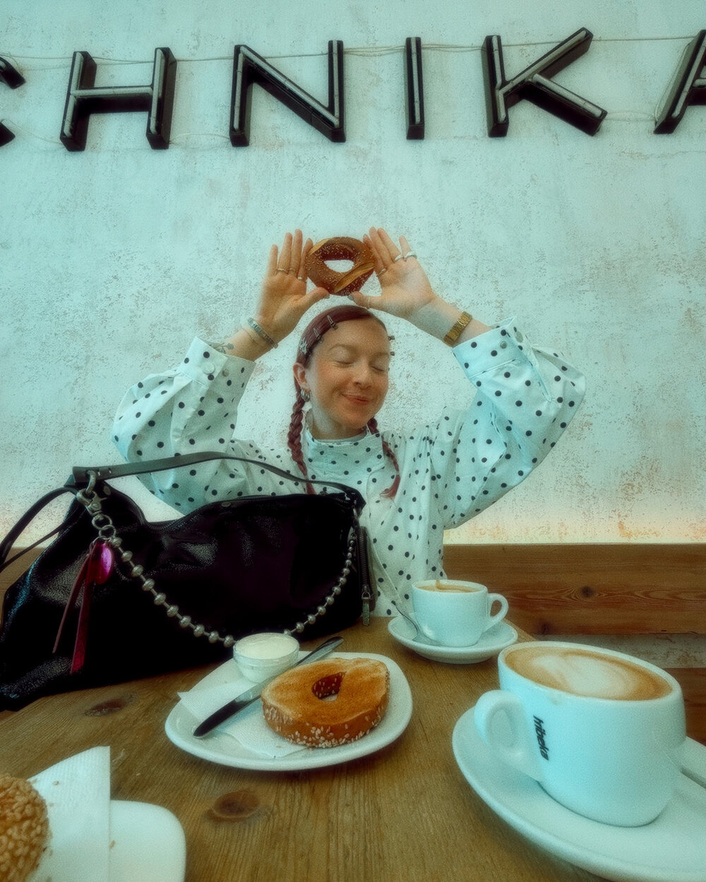 Eine Frau in einer gepunkteten Bluse hält ein Gebäckstück über ihren Kopf und sitzt an einem Tisch mit Kaffee und einem Donut, neben ihr steht ihre elegante schwarze INA KENT-Handtasche.