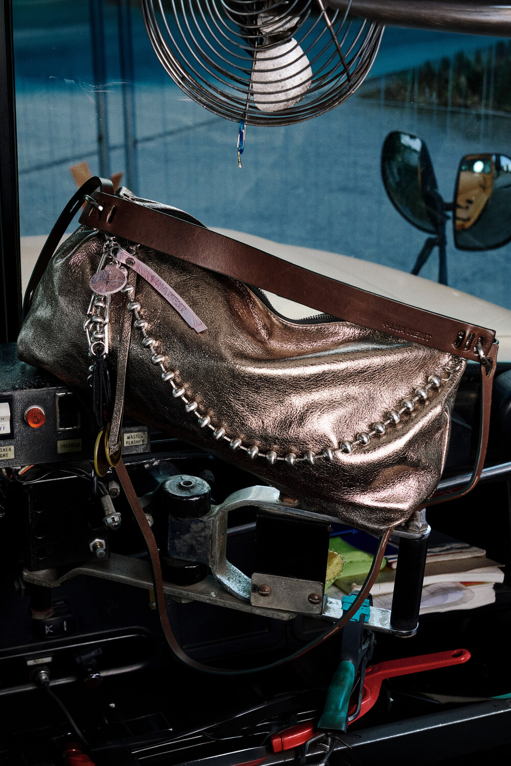An INA KENT metallic handbag with a brown strap and silver zipper hangs inside a vehicle, surrounded by various equipment, wires, and a fan.