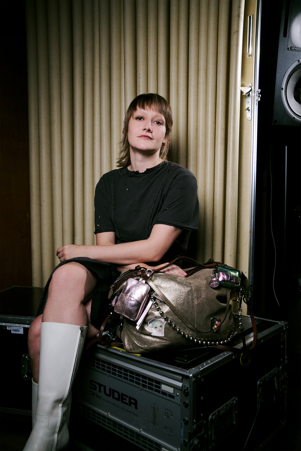 A person with short hair sits on equipment next to an INA KENT gold bag, wearing a dark dress and white knee-high boots, with a neutral expression.