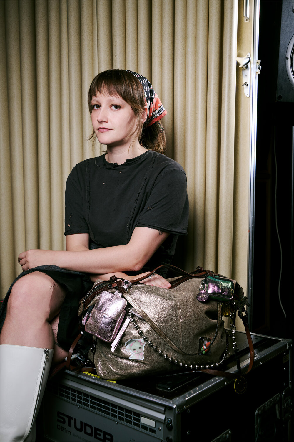 A woman in a black dress and white boots sits beside an INA KENT metallic handbag adorned with assorted keychains, in front of beige curtains and audio equipment.