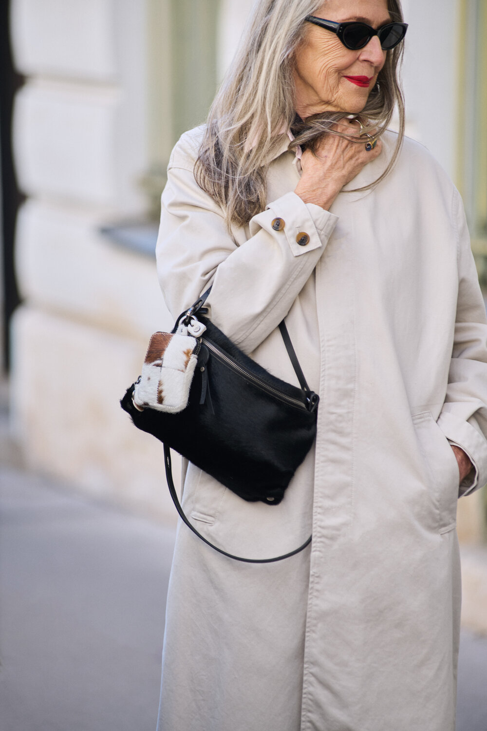 Woman in a beige trench coat and sunglasses stands outdoors, holding her collar. She carries an INA KENT black crossbody bag with a small white and brown pouch attached.