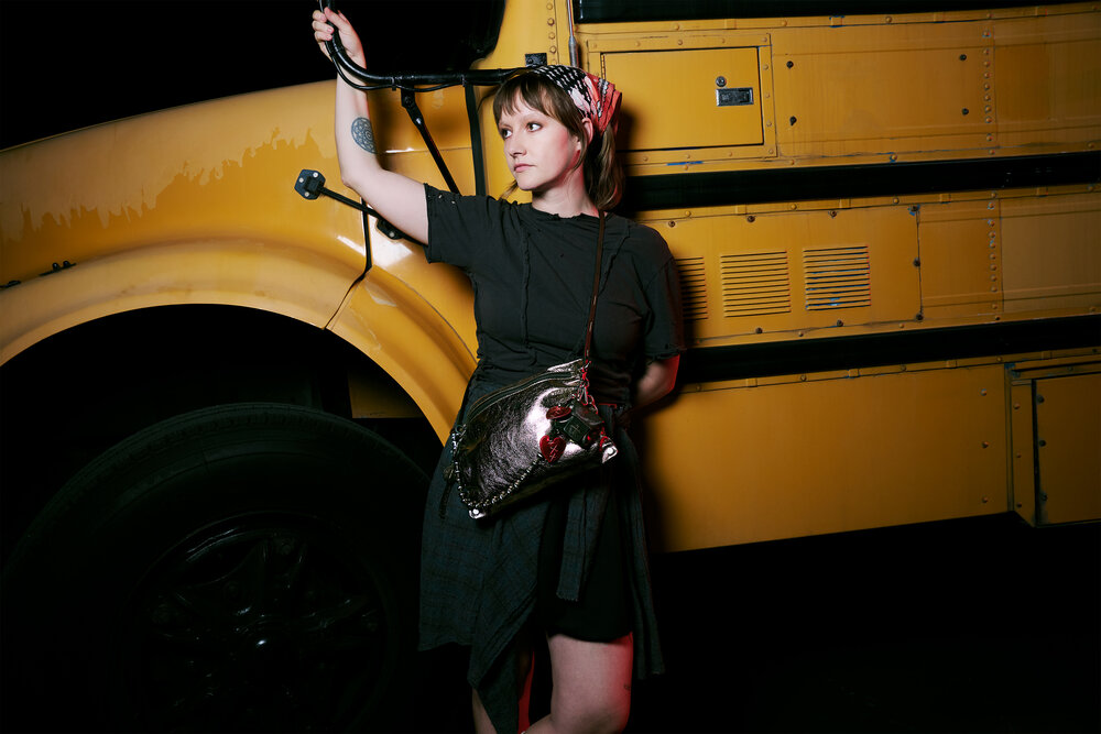 Person with short hair and headband leans against a yellow school bus, wearing a dark outfit and carrying a shiny INA KENT handbag shaped like a fish.