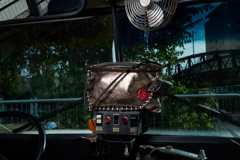 A metallic INA KENT handbag with a heart keychain sits atop a control panel inside a vehicle, with a fan, windshield, and outdoor scenery visible in the background.