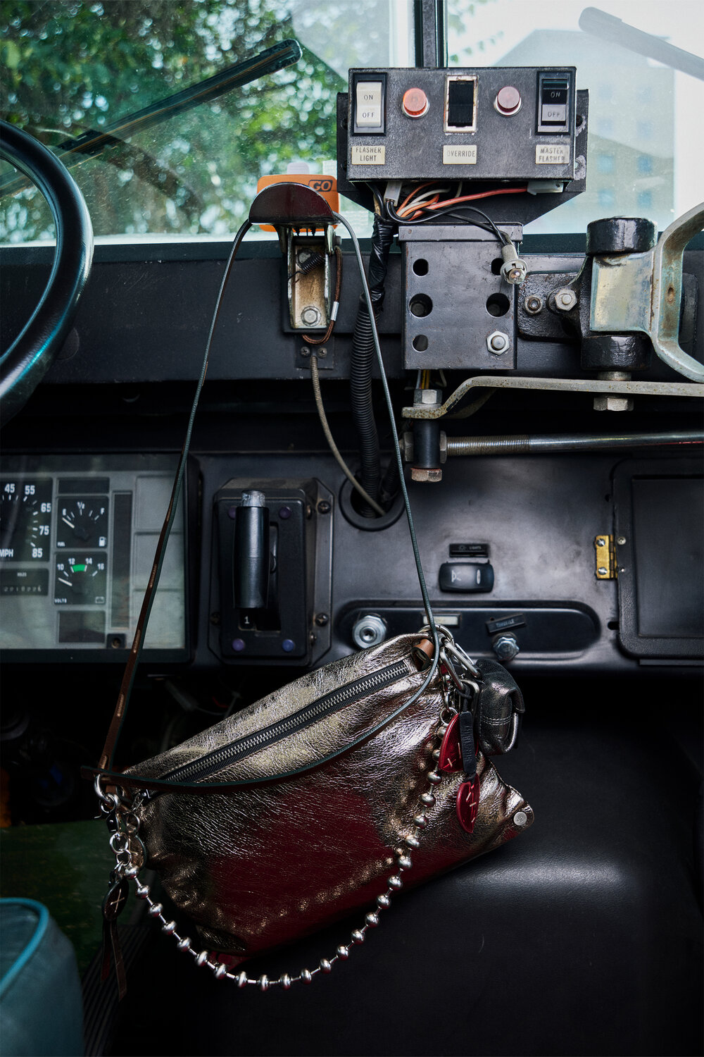 A metallic INA KENT handbag with a studded strap hangs from a control panel inside a vehicle, with dashboard instruments and windshield visible in the background.