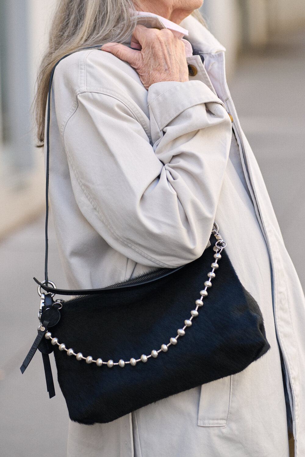 An older woman wearing a light-colored coat carries an INA KENT black handbag with a pearl-like beaded strap over her shoulder.
