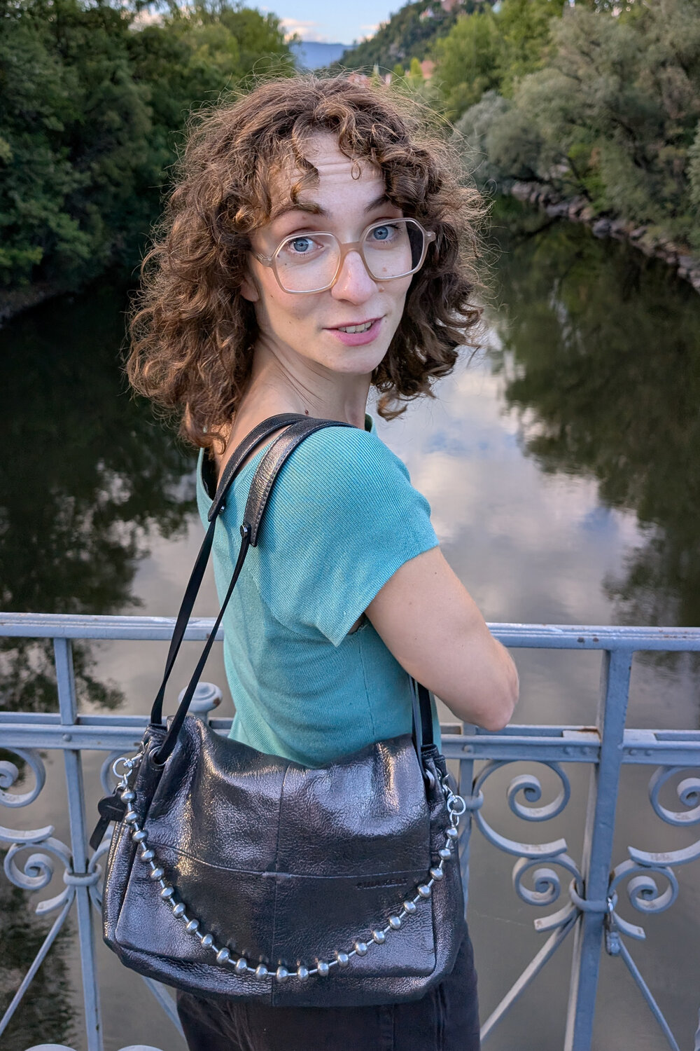 Eine Person mit lockigem Haar und Brille, die ein blaugrünes Hemd trägt und eine metallische INA KENT-Umhängetasche bei sich hat, steht auf einer Brücke über einem Fluss.