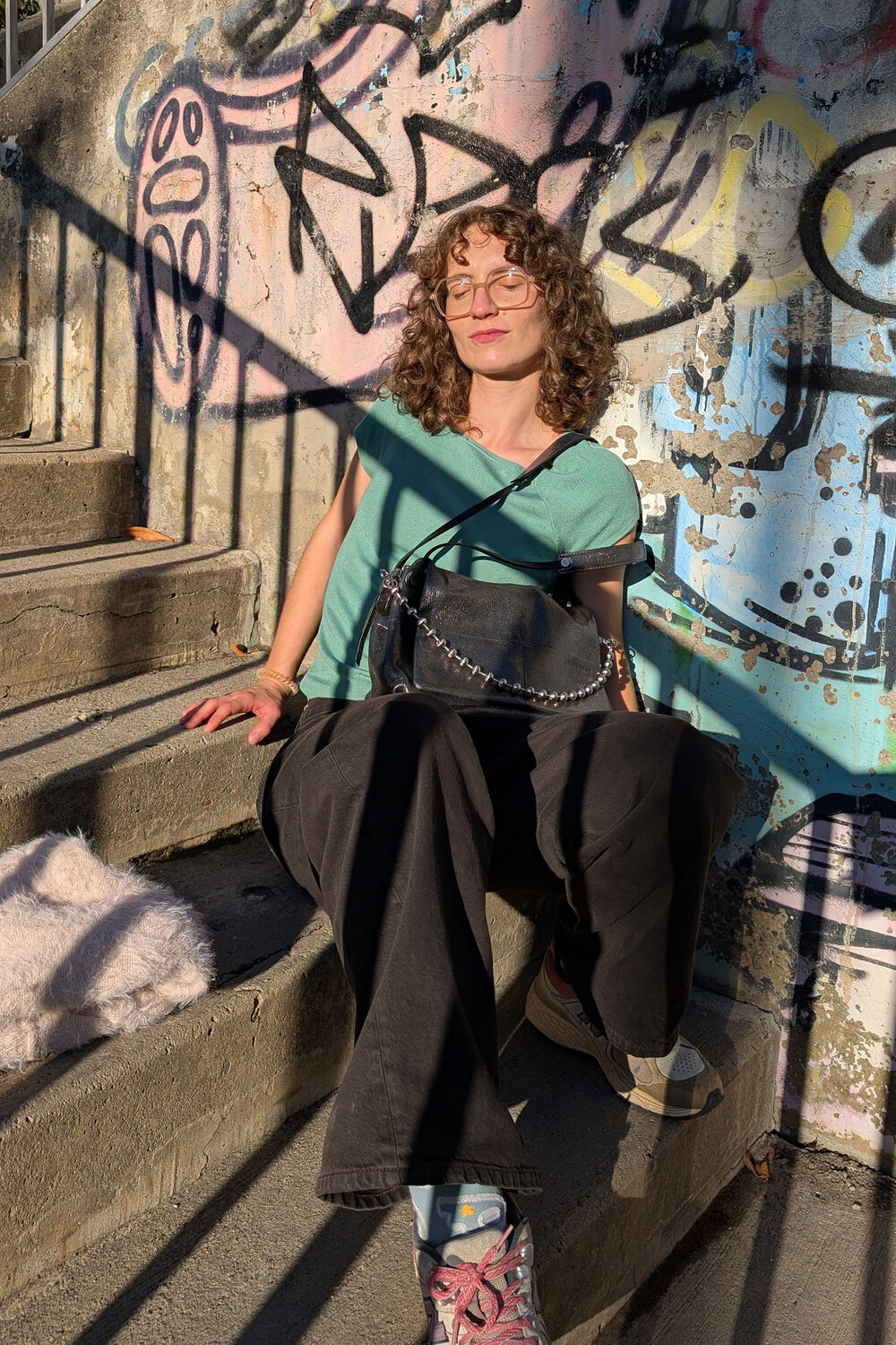 Eine Person mit lockigem Haar und Brille, die ein Accessoire von INA KENT trägt, sitzt auf einer Betonstufe vor einer mit Graffiti übersäten Wand und hat in der Sonne die Augen geschlossen.