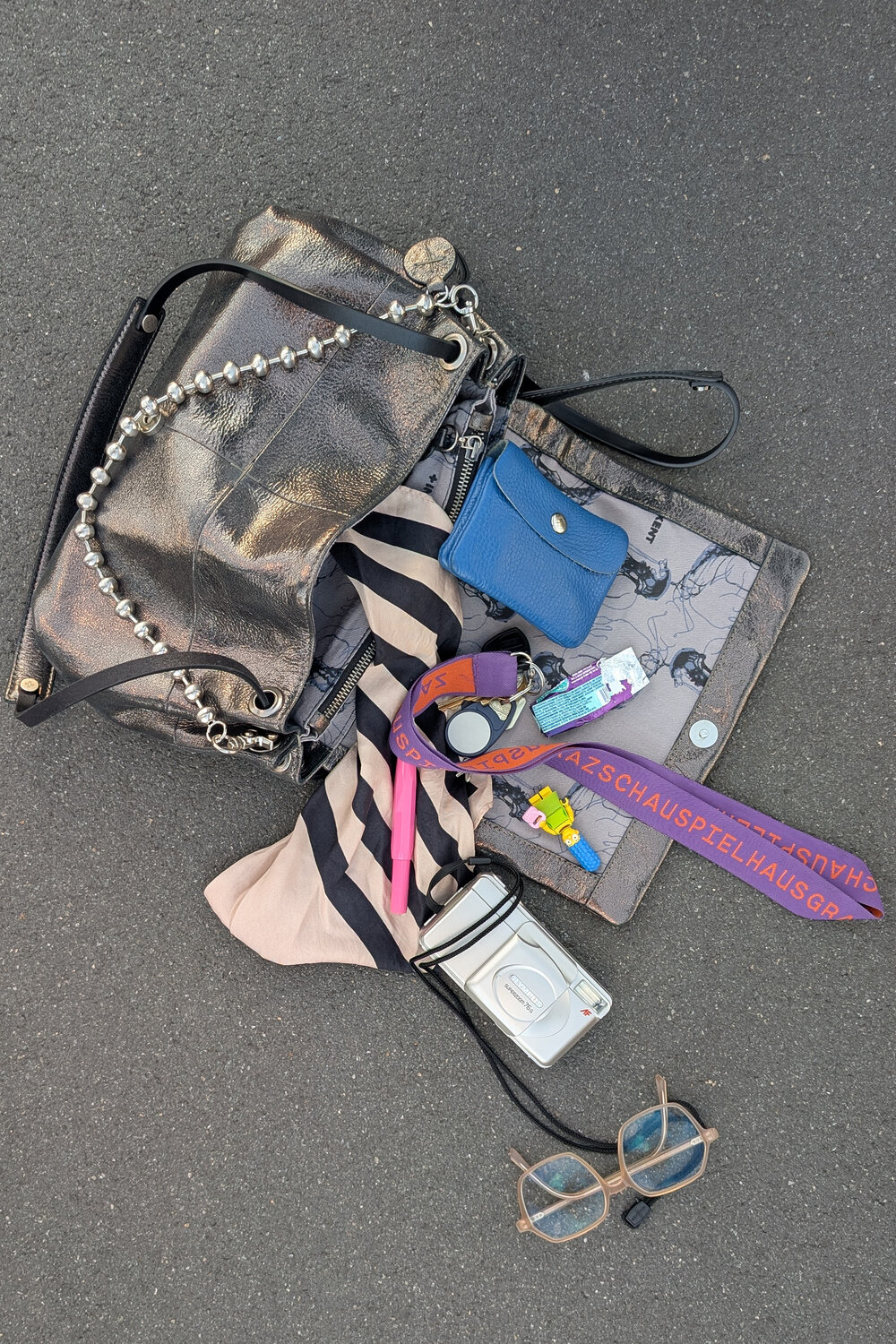 Eine offene Metallhandtasche der Marke INA KENT auf Asphalt, aus der der Inhalt herausgefallen ist, darunter eine blaue Geldbörse, ein lila Schlüsselband, eine Kamera, eine Brille und ein gestreifter Schal.