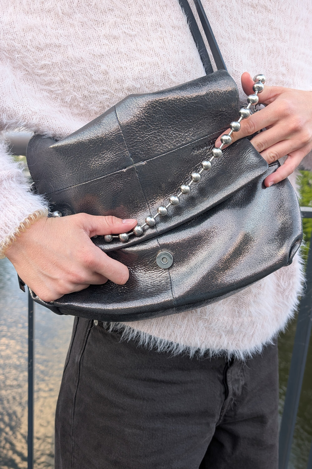 Eine metallisch graue INA KENT-Umhängetasche mit einem Kettenriemen mit Perlenverzierung, die von einer Person in einem flauschigen Pullover getragen wird.
