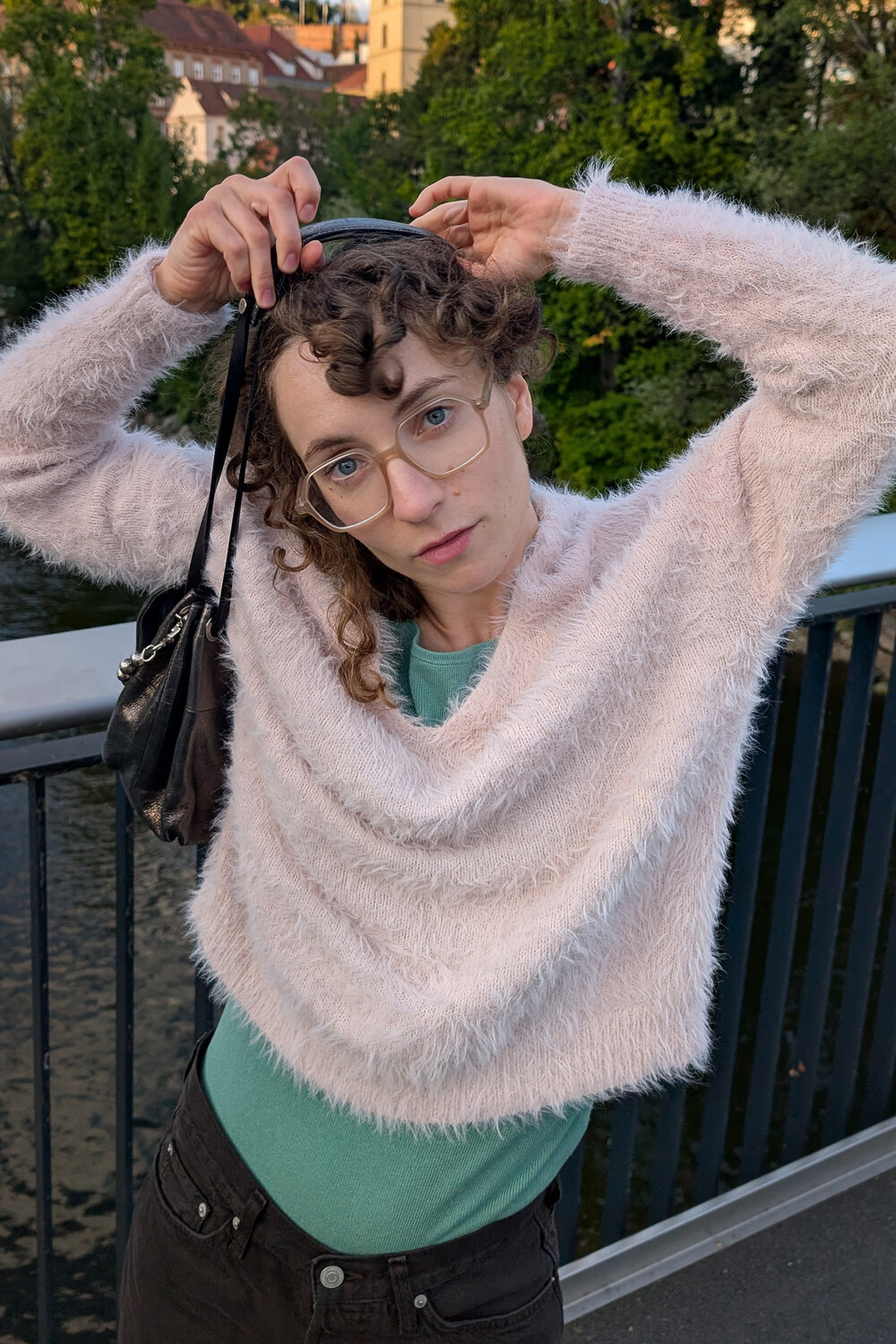 Eine Person mit lockigem Haar und Brille steht auf einer Brücke und trägt einen flauschigen rosa Pullover und ein grünes INA KENT-Shirt.