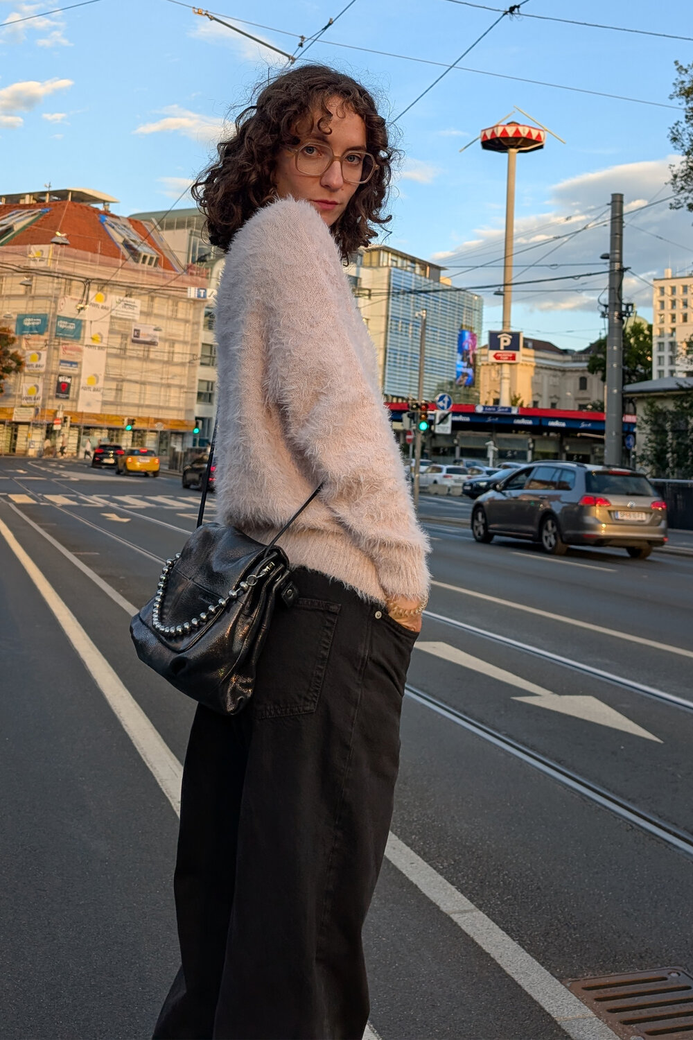 Eine Person mit lockigem Haar, die einen flauschigen Pullover und eine schwarze Hose trägt, steht auf einer Straße in der Stadt und hält eine INA KENT-Umhängetasche in der Hand.