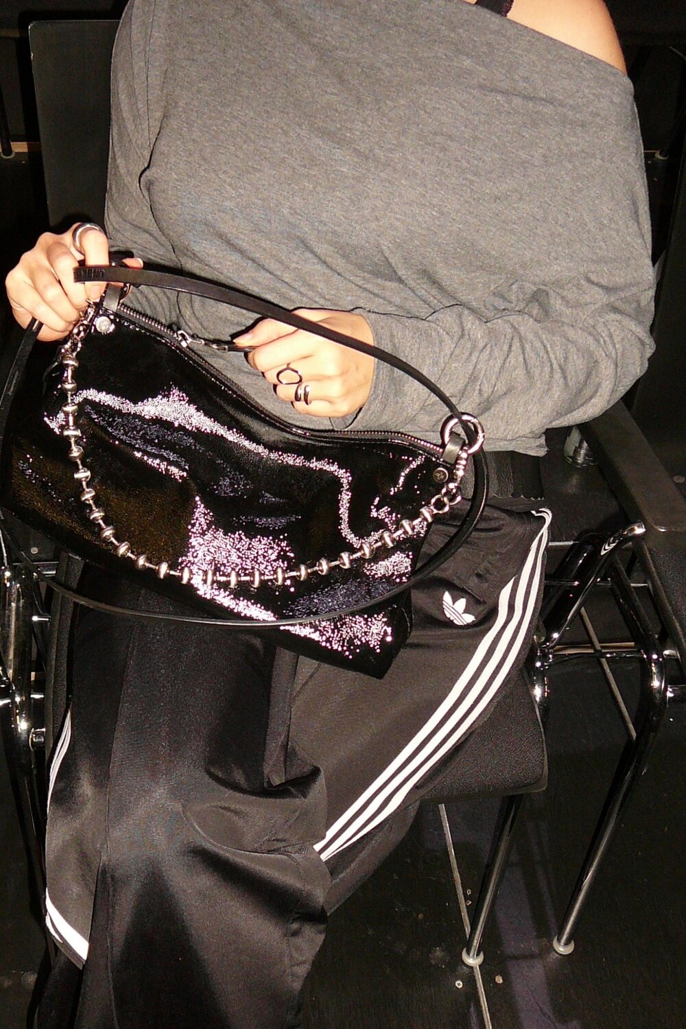 Eine Person in einem grauen, schulterfreien Oberteil und einer Adidas-Jogginghose hält eine glänzende schwarze INA KENT-Handtasche mit einer silbernen Kette in der Hand.
