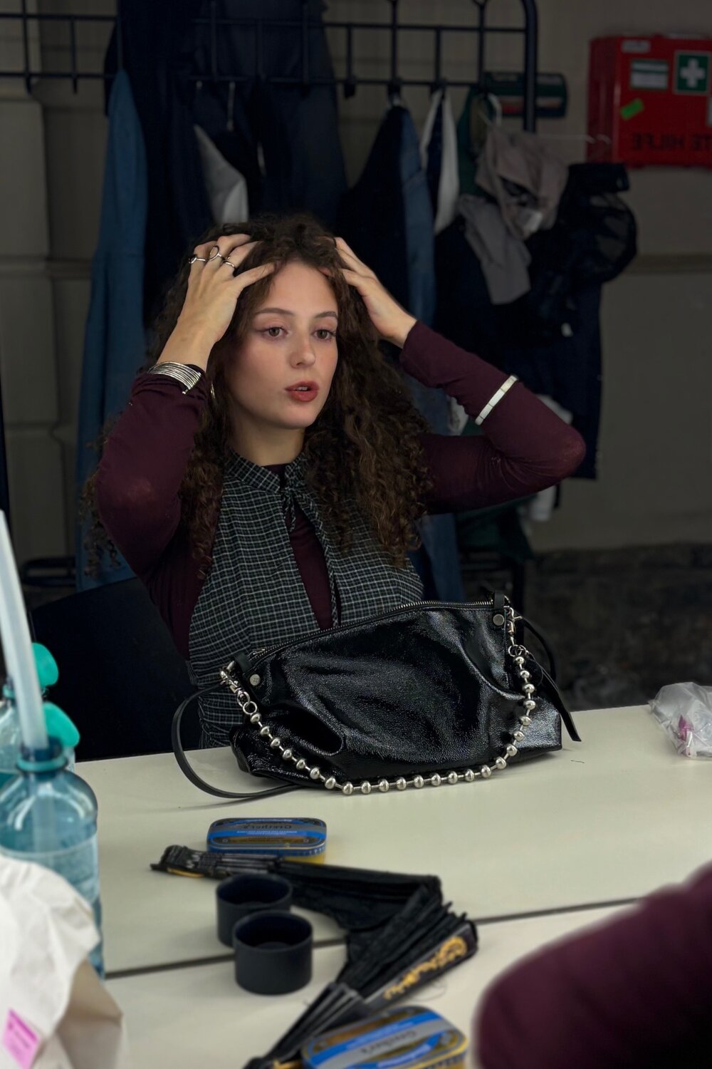 Eine Frau mit lockigem Haar sitzt an einem Tisch und richtet ihre Haare vor einem Spiegel, während eine INA KENT-Handtasche vor ihr liegt.