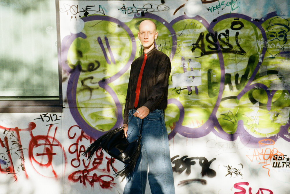 Eine Person mit rasiertem Kopf, die ein dunkles Hemd, eine rote Krawatte und eine Jeans mit weitem Bein trägt und eine schwarze Tasche mit Fransen vor einer mit Graffiti übersäten Wand in der Hand hält.