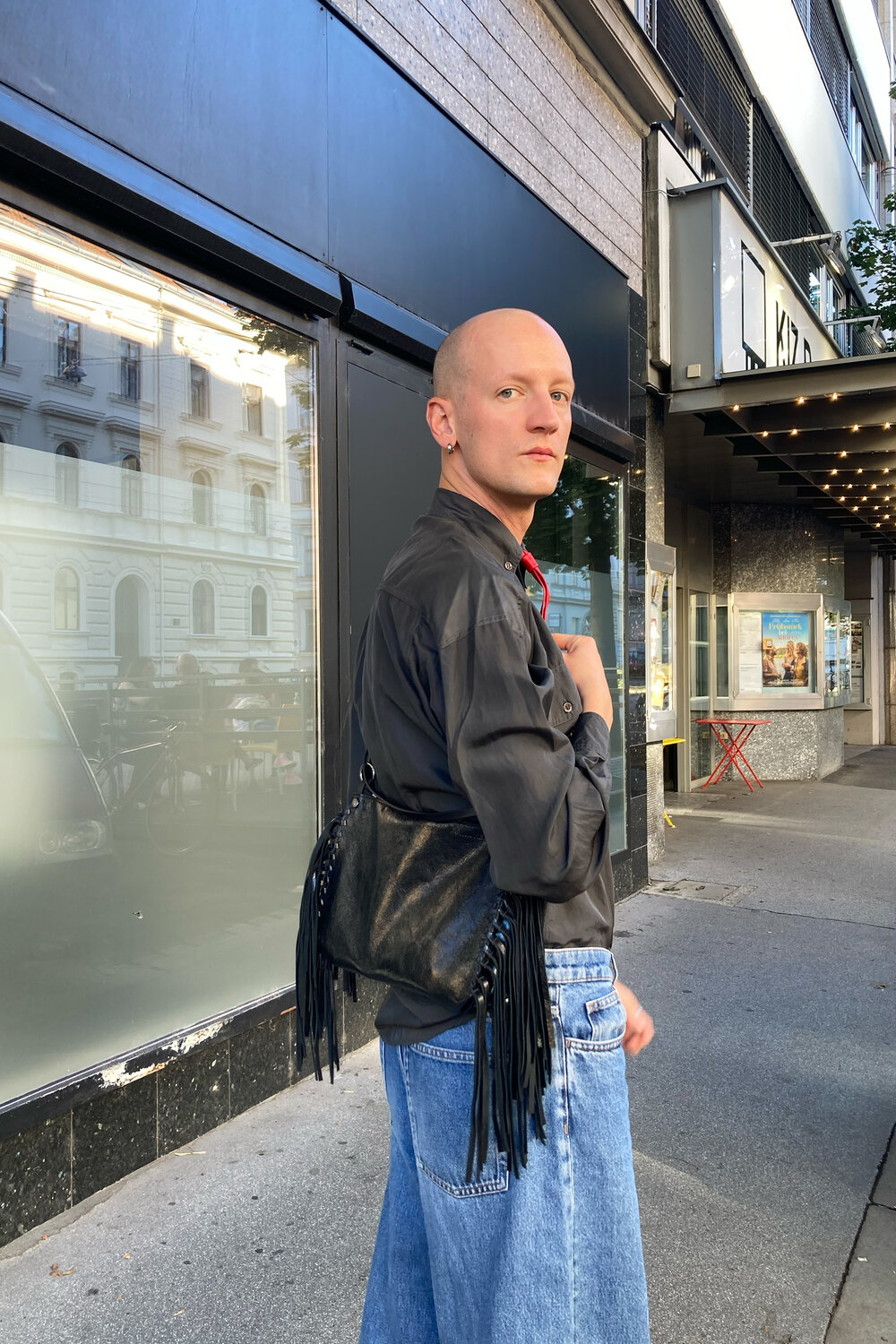 Eine Person mit rasiertem Kopf, die ein dunkles Hemd und locker sitzende Jeans trägt und eine schwarze Schultertasche mit Fransen auf einem Bürgersteig in der Stadt bei sich hat.