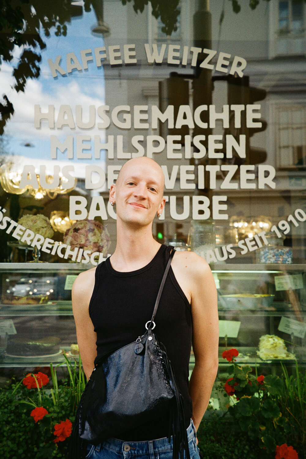 Eine Person, die ein schwarzes Tanktop und eine Umhängetasche trägt, steht vor dem Fenster eines Cafés, auf dessen Scheibe ein deutscher Text zu lesen ist.