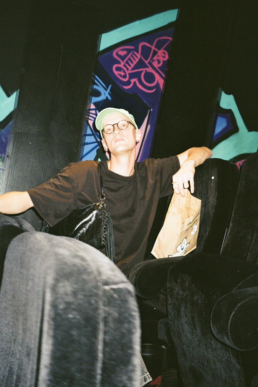 Eine Person mit Brille und grüner Baseballkappe, die auf einem Theatersessel sitzt und eine Papiertüte in der Hand hält.