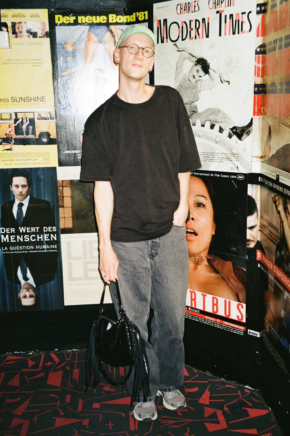 Eine Person in einem schwarzen T-Shirt, grauen Jeans und einer grünen Mütze steht vor einer Wand, die mit Filmplakaten bedeckt ist, und hält eine schwarze Handtasche mit Fransen in der Hand.