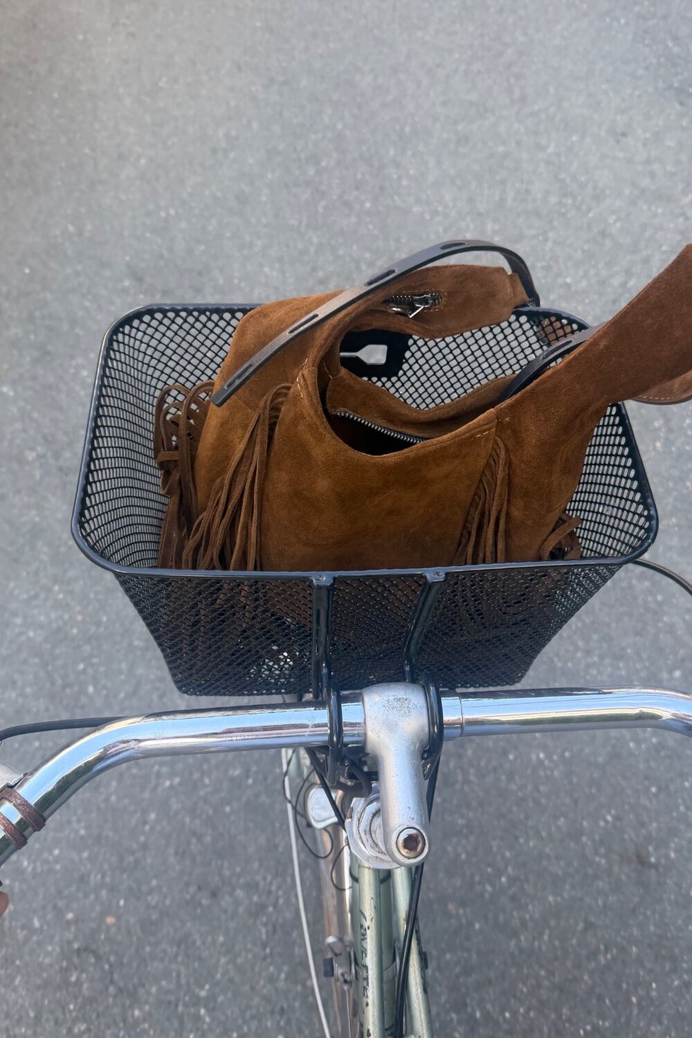 Eine braune Wildlederhandtasche mit Fransen von INA KENT, die in einem schwarzen Fahrradkorb aus Metall liegt.