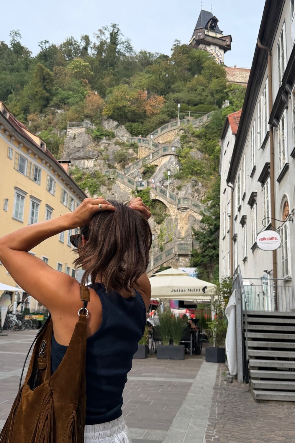 Eine Person mit einer INA KENT-Tasche steht auf einer Straße in der Stadt und blickt auf eine steile, gewundene Treppe, die einen felsigen Hang hinauf zu einem Uhrturm führt.