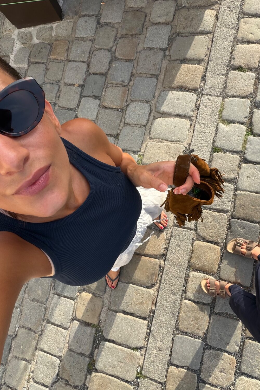 Eine Person in einem schwarzen Tanktop und mit Sonnenbrille macht auf einer Kopfsteinpflasterstraße ein Selfie, während sie eine braune Tasche mit Fransen von INA KENT in der Hand hält.