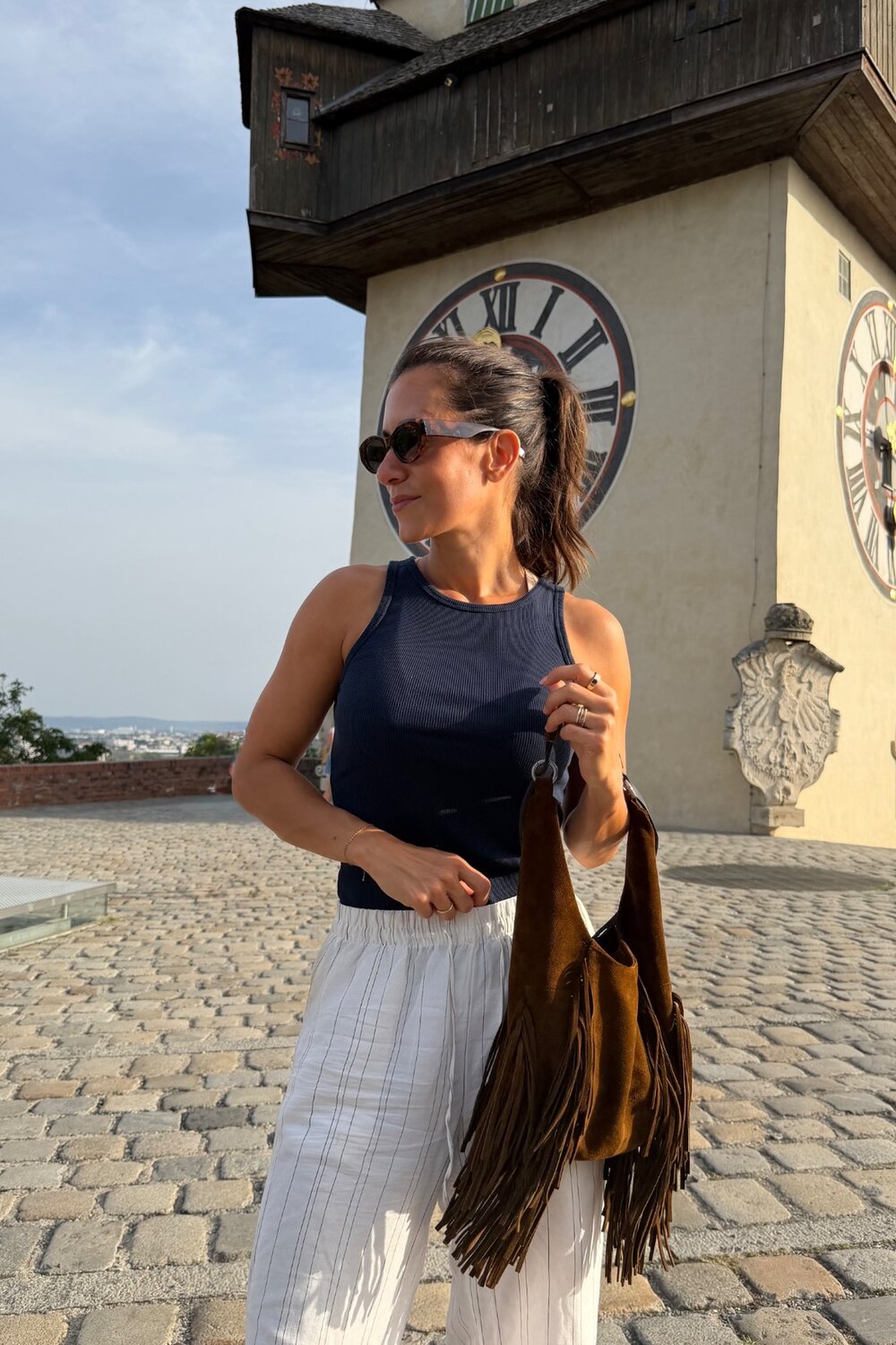 Eine Frau in einem dunklen Tanktop und einer gestreiften Hose steht vor dem Grazer Uhrturm und hält eine braune Wildleder-Tasche mit Fransen von INA KENT in der Hand.