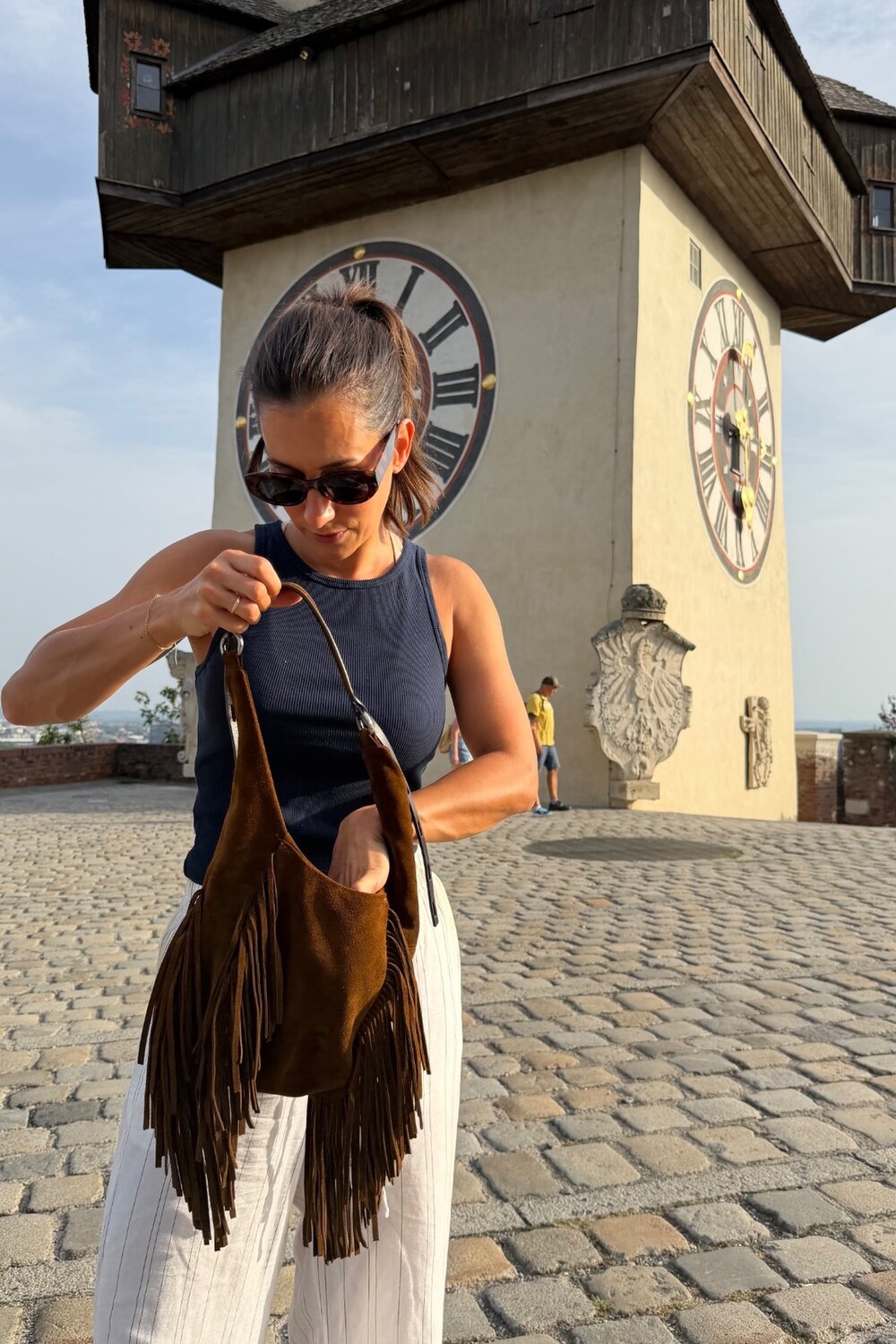 Eine Frau mit Sonnenbrille und einem marineblauen Tanktop greift in eine braune Wildlederhandtasche mit Fransen von INA KENT, im Hintergrund ist der Grazer Uhrturm zu sehen.