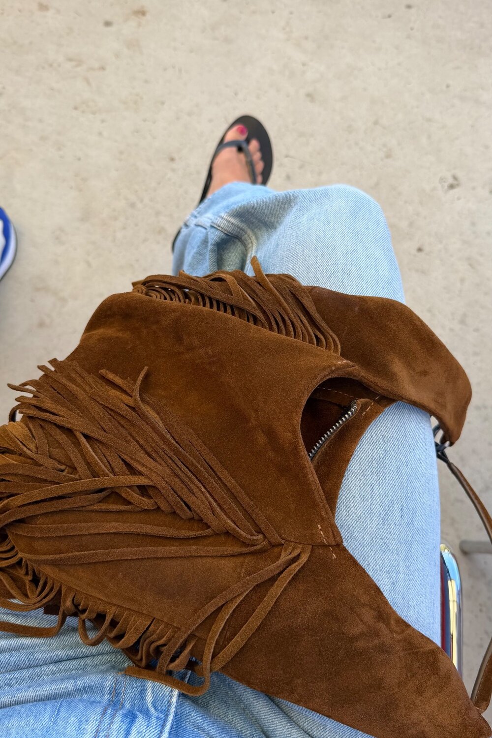 Eine braune Wildlederhandtasche mit Fransen von INA KENT, die auf dem Schoß einer Person liegt, die eine hellgewaschene Jeans trägt.
