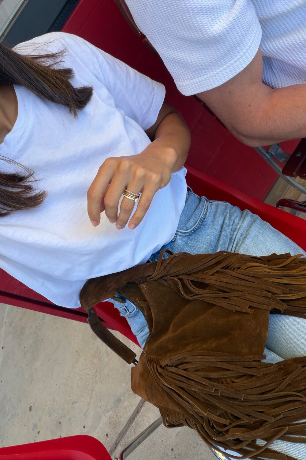 Eine Frau in einem weißen T-Shirt und Jeans sitzt auf einem roten Stuhl, auf ihrem Schoß liegt eine braune Wildledertasche mit Fransen von INA KENT.