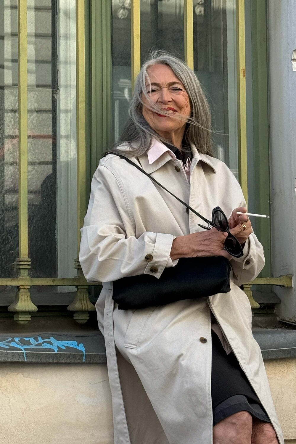 Older woman with long gray hair in a beige coat stands outside in front of a window, holding sunglasses and a cigarette, with a slight smile and wind blowing her hair.