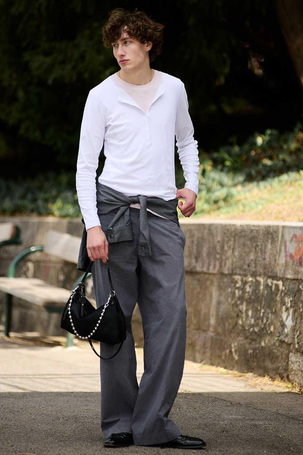 A person stands outdoors wearing a white long-sleeve shirt, gray wide-leg pants, and a sweatshirt tied at the waist, holding a black handbag.