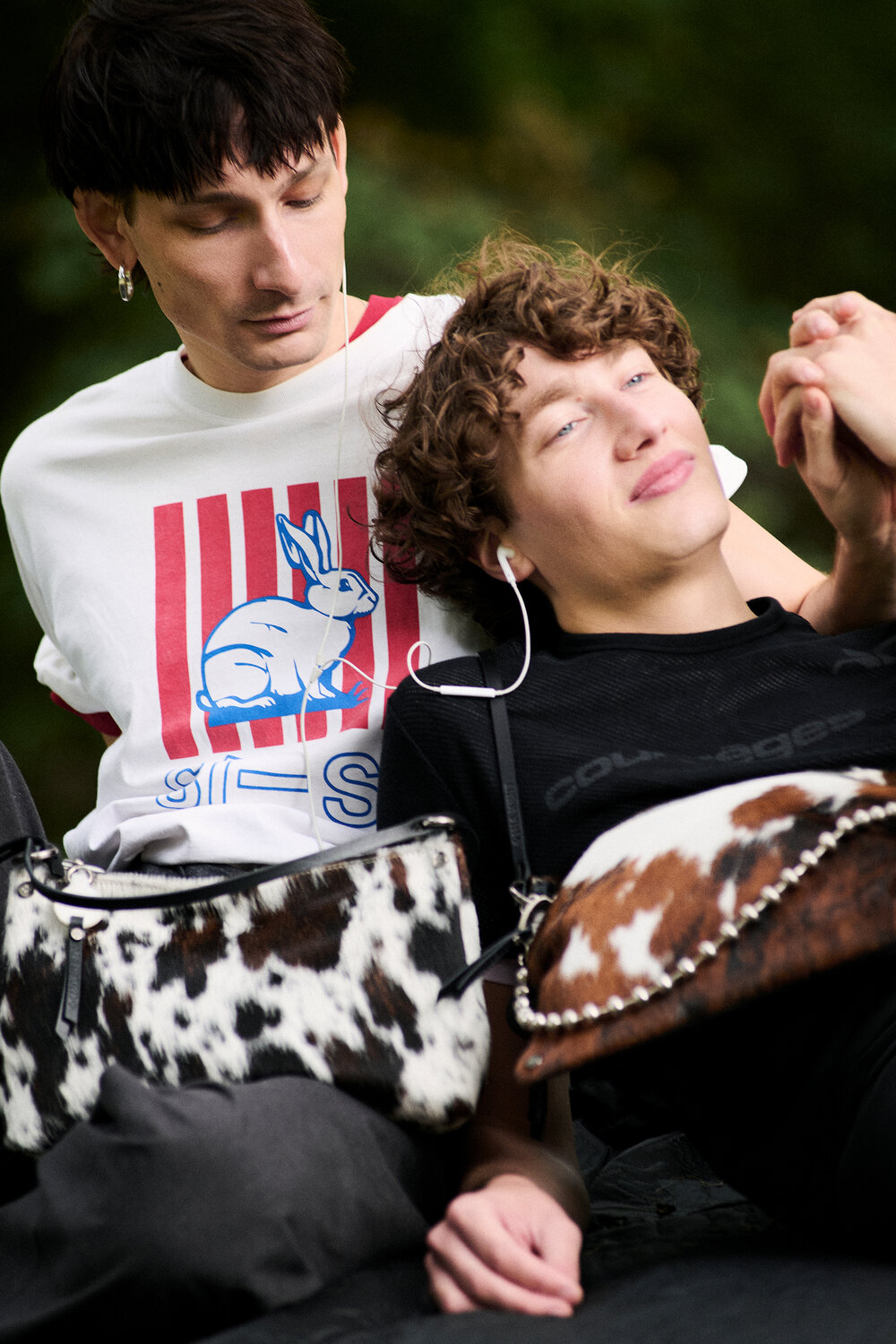 Zwei junge Männer sitzen eng beieinander; einer trägt ein T-Shirt mit einem weißen Kaninchenaufdruck, der andere liegt auf seinem Schoß und hört Musik. Beide tragen INA KENT-Taschen mit Rindsledermuster. Grüner Outdoor-Hintergrund.