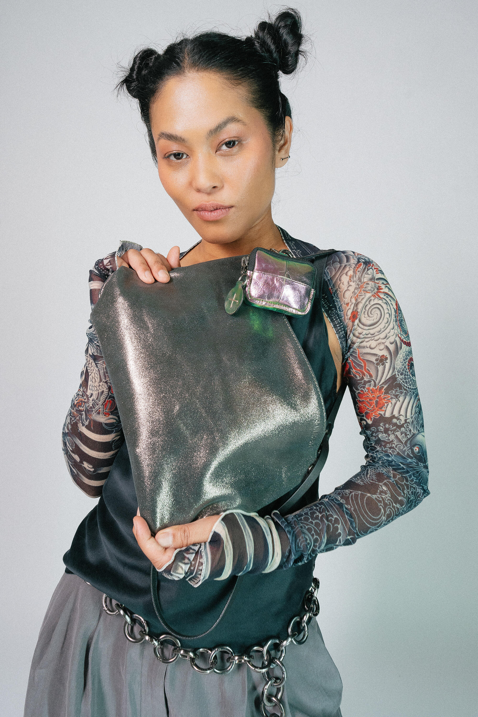 Eine Person mit frisiertem Haar hält eine große metallische INA KENT-Handtasche dicht vor der Brust. Sie trägt ein Oberteil mit Spitzenmuster und langen Ärmeln sowie einen Kettengürtel.